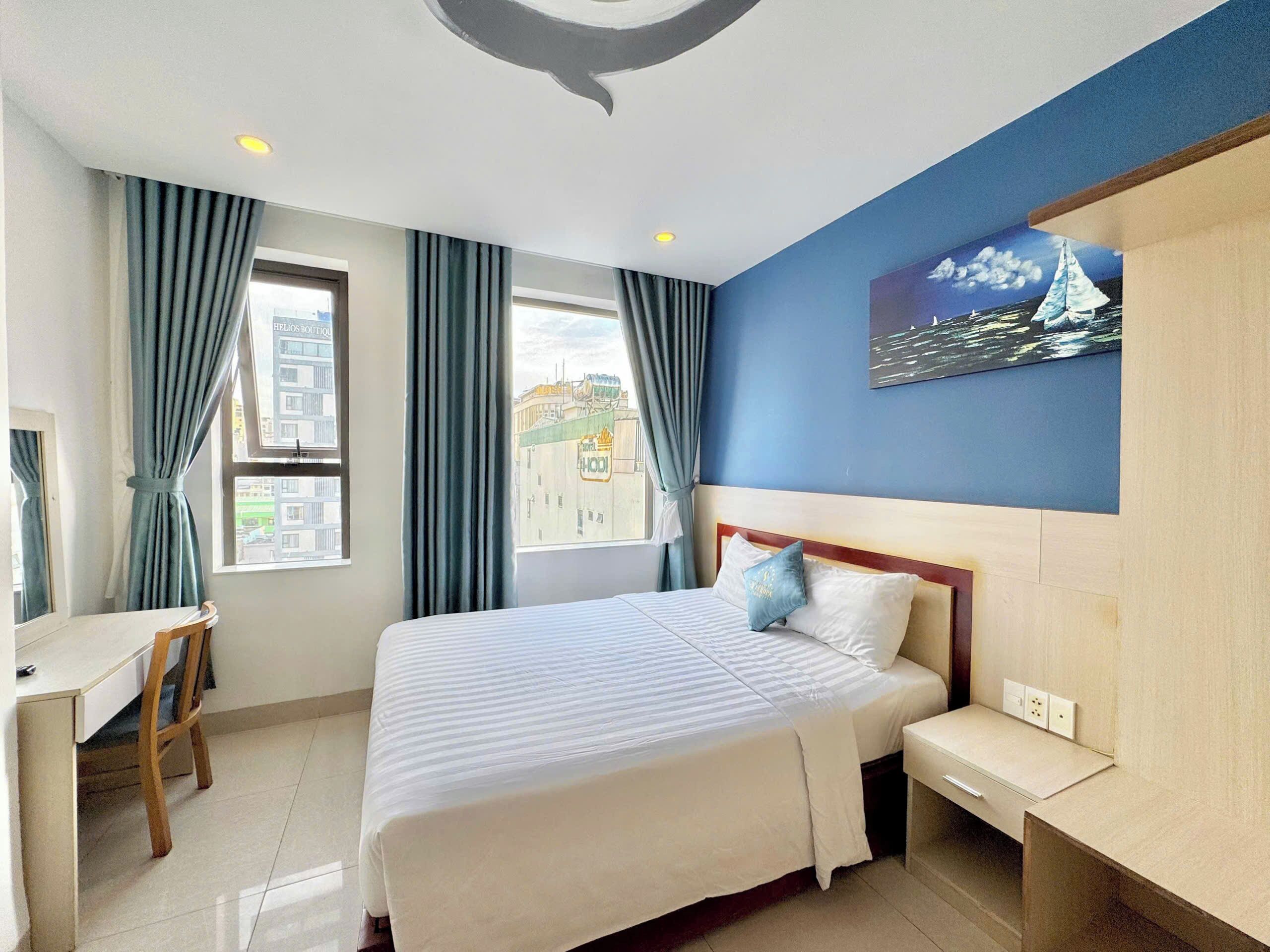 A hotel room with a bed, a desk, and a chair. There are windows with curtains, a painting on the wall, and a view of buildings outside.