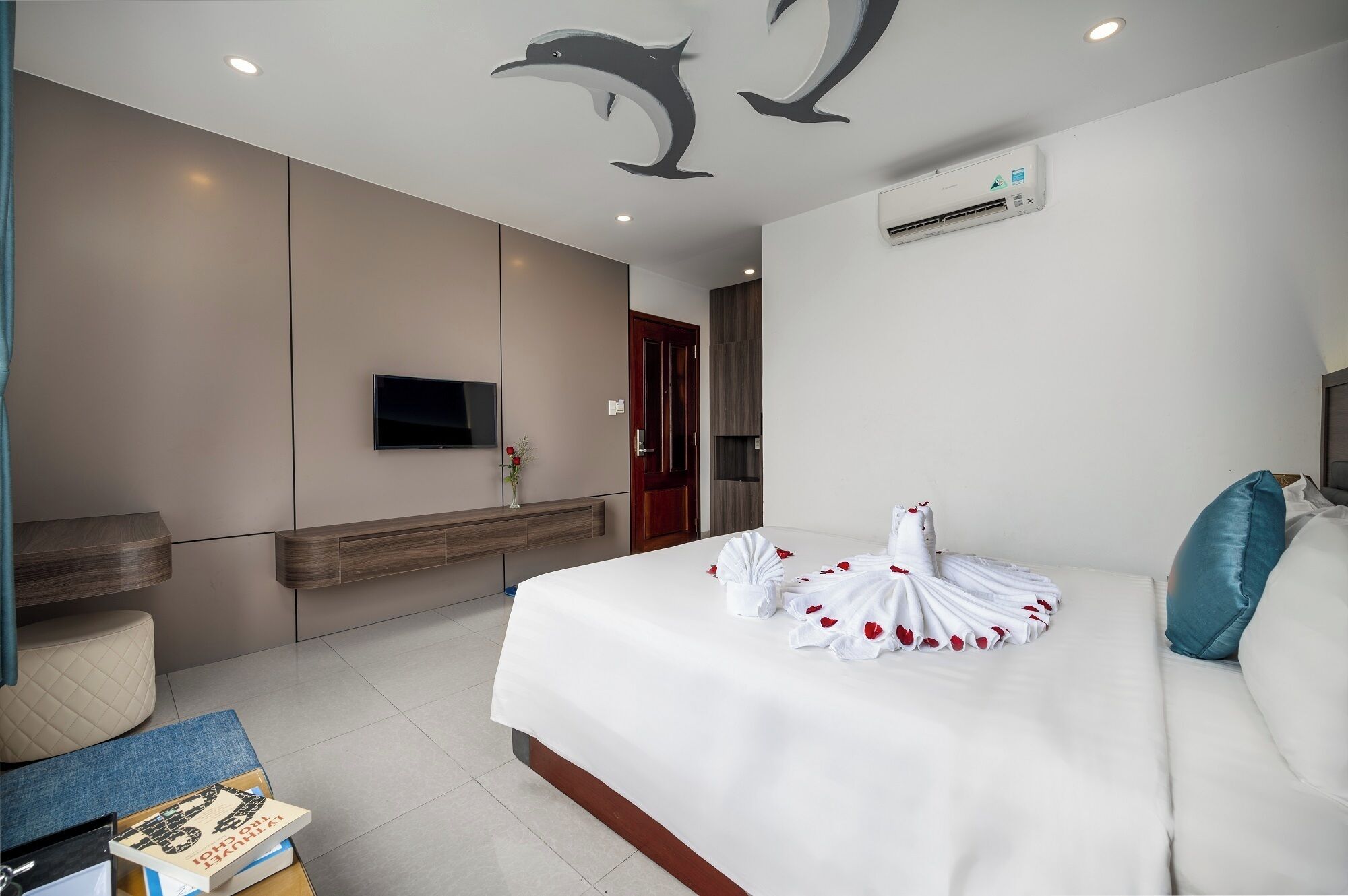 A modern hotel room with a large bed, a flat-screen TV, a wall-mounted air conditioner, and a ceiling with dolphin artwork.