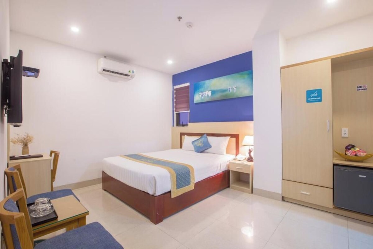 Quarto de hotel com cama, mesa de cabeceira, cadeira, escrivaninha e televisão na parede.
