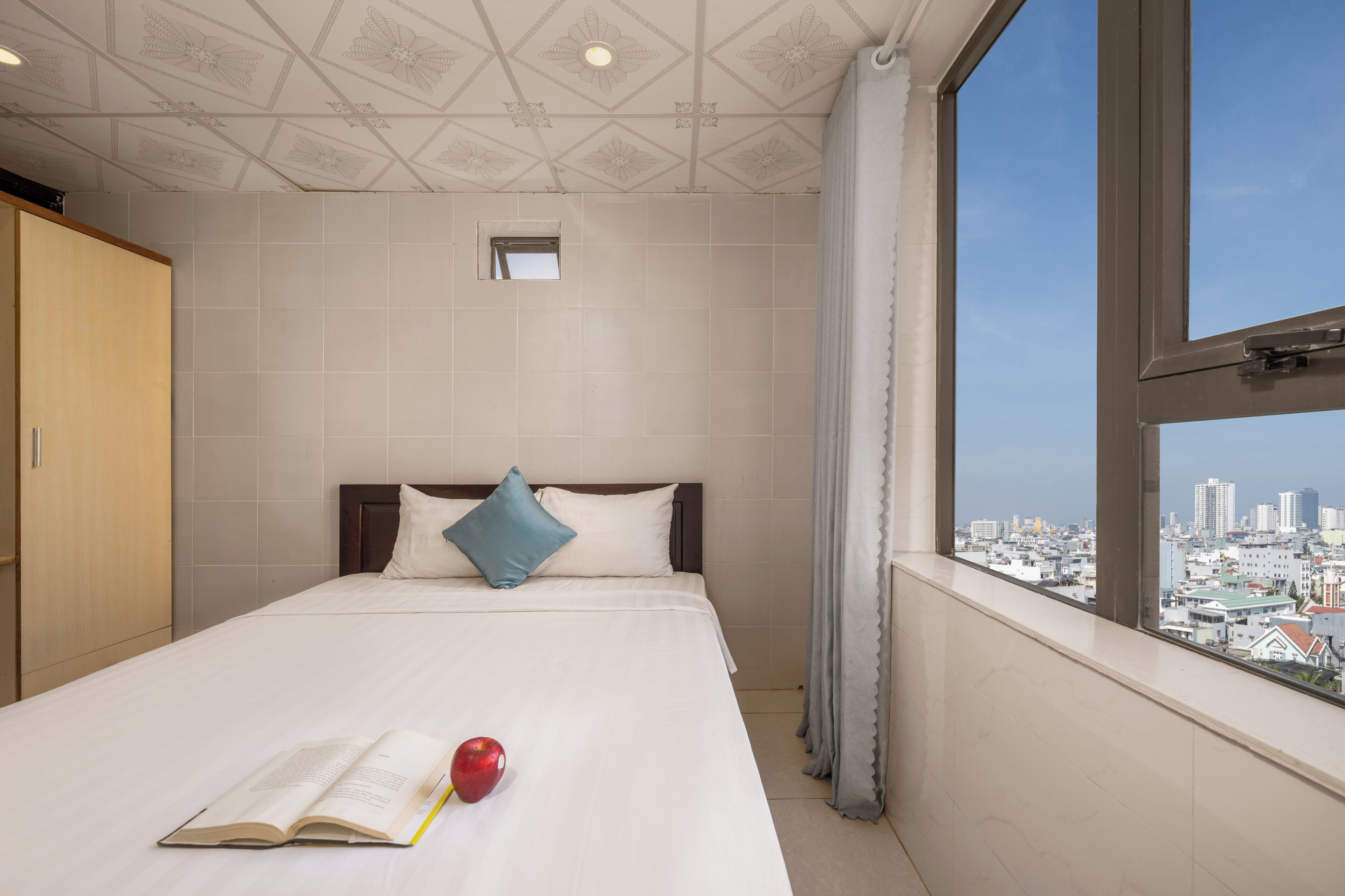 A hotel room with a bed, a window with a city view, a wardrobe, and a small window above the bed.