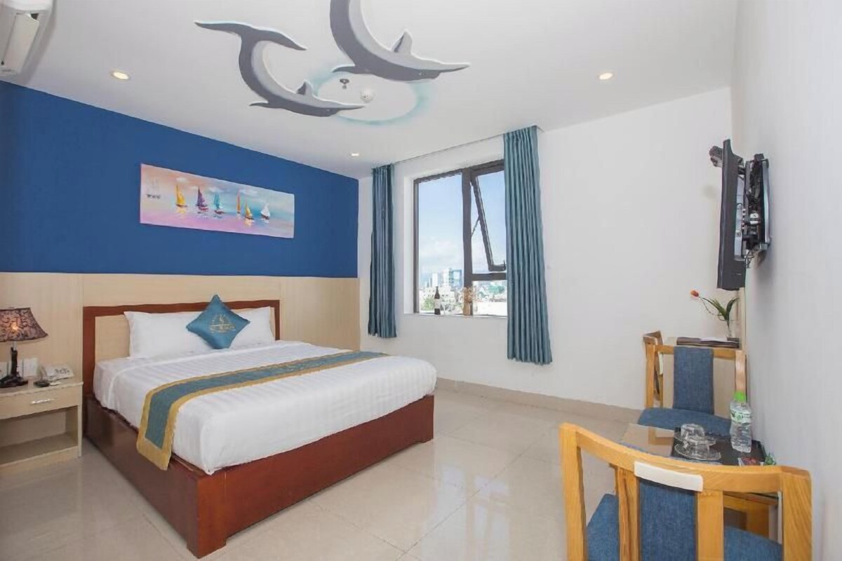 Quarto de hotel com cama, escrivaninha, cadeira, janela com cortinas e um quadro na parede.