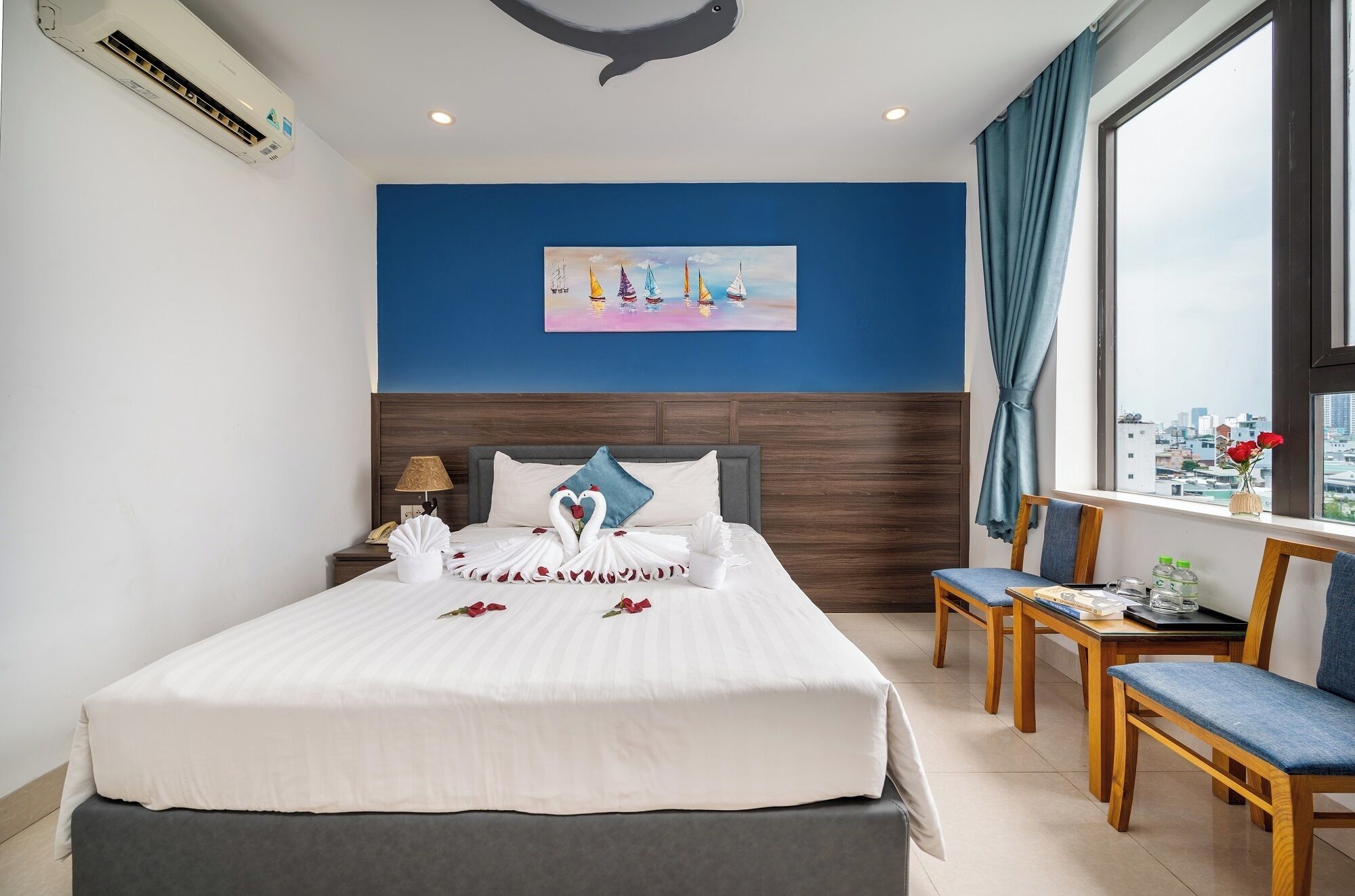 A hotel room with a bed, a desk, and a chair. There is a painting on the wall and a window with a view of the city.
