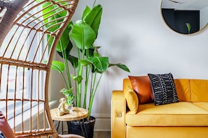 Ein Zimmer mit einem gelben Sofa, einem runden Spiegel, einem Hängekorbstuhl und einer großen Grünpflanze.