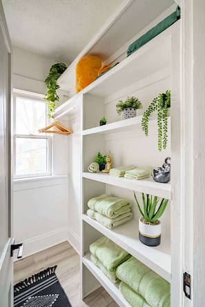 A white interior with a wooden floor, a hanging plant, a window, and a shelf with folded towels. - Short North & Downtown & OSU | Near High Street | Free Parking | 5,800+ Reviews (Columbus)