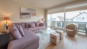 A modern living room with a sectional sofa, glass coffee table, ottomans, and a wicker chair. - Apartment in Duinbergen Near Sailing School (Knokke-Heist)