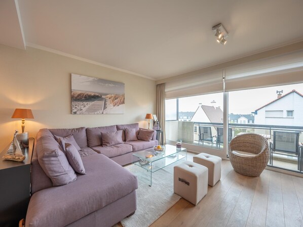 A modern living room with a sectional sofa, glass coffee table, ottomans, and a wicker chair. - Apartment in Duinbergen Near Sailing School (Knokke-Heist)