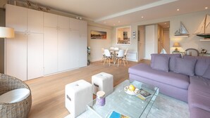A modern living room with a purple sofa, a glass coffee table, and a wicker chair. There is a dining area with a table and chairs, and a kitchen with white cabinets. - Apartment in Duinbergen Near Sailing School (Knokke-Heist)