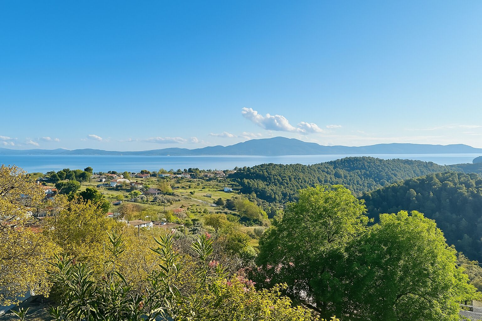 Vista panorámica de un pueblo costero con cielo despejado, abundante vegetación y una cadena montañosa distante.