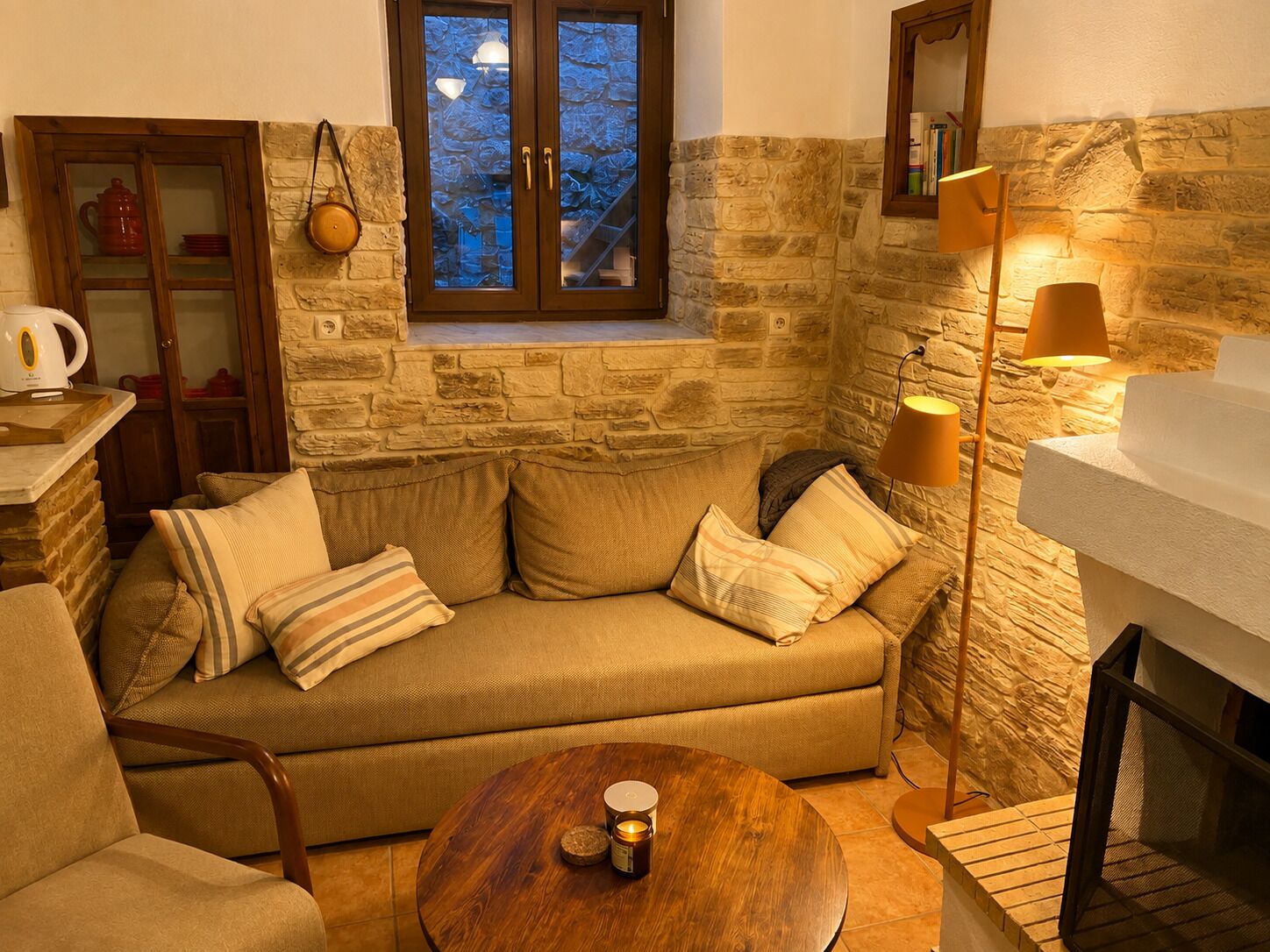 Una sala de estar acogedora con una pared de piedra, un mueble de madera, un sofá con cojines, una mesa redonda de madera y una lámpara de pie.