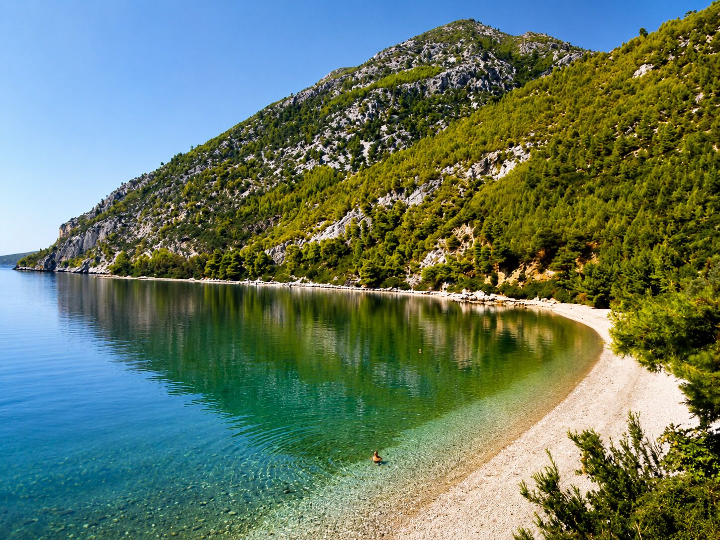 Un lago transparente con una playa arenosa y una montaña boscosa al fondo.