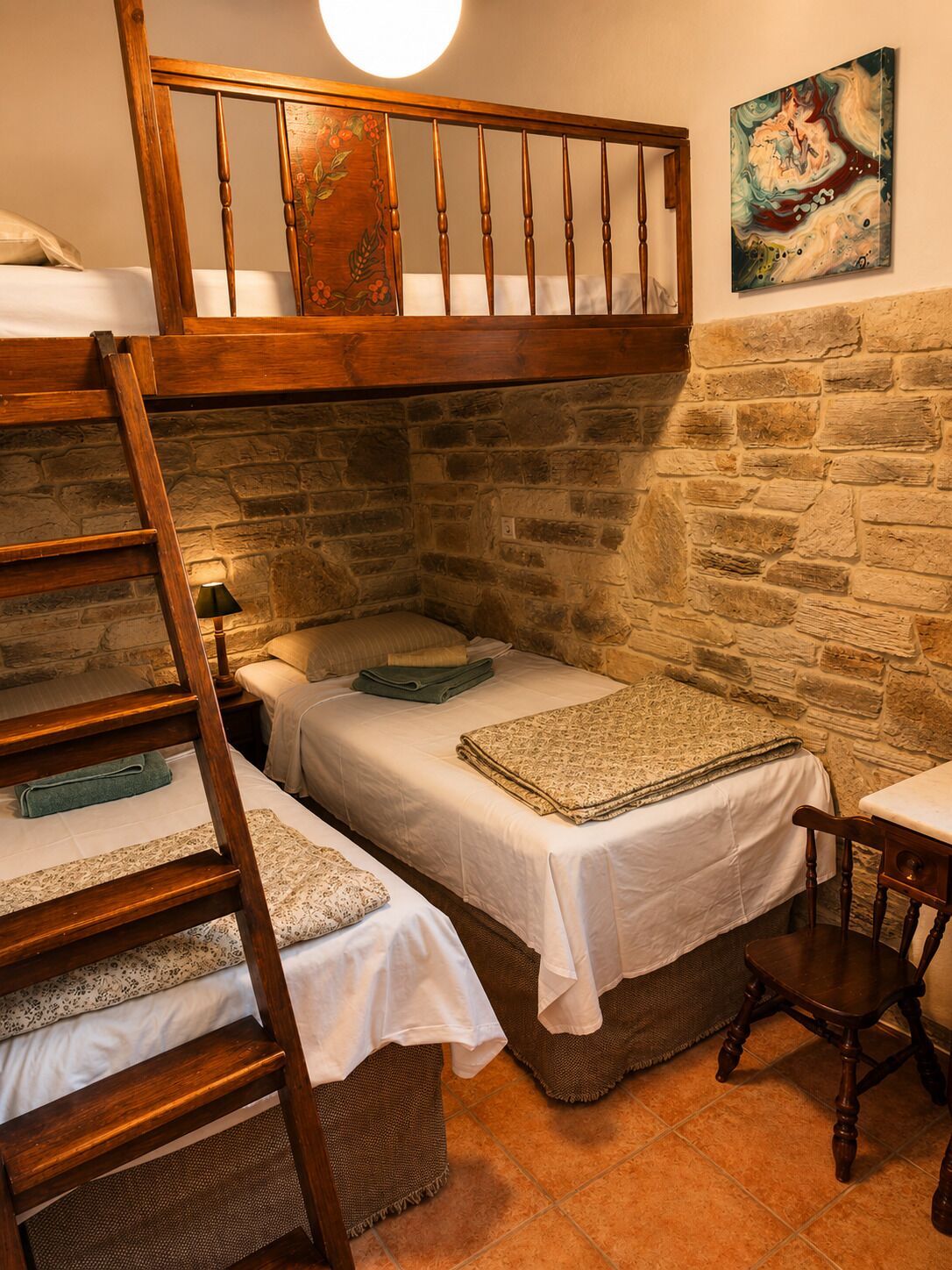 Una habitación rústica con cama en altillo, dos camas bajas, escalera de madera, pared de piedra, un cuadro y una pequeña mesa.
