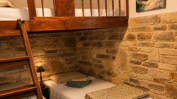 Una habitación rústica con cama en altillo, dos camas bajas, escalera de madera, pared de piedra, un cuadro y una pequeña mesa.