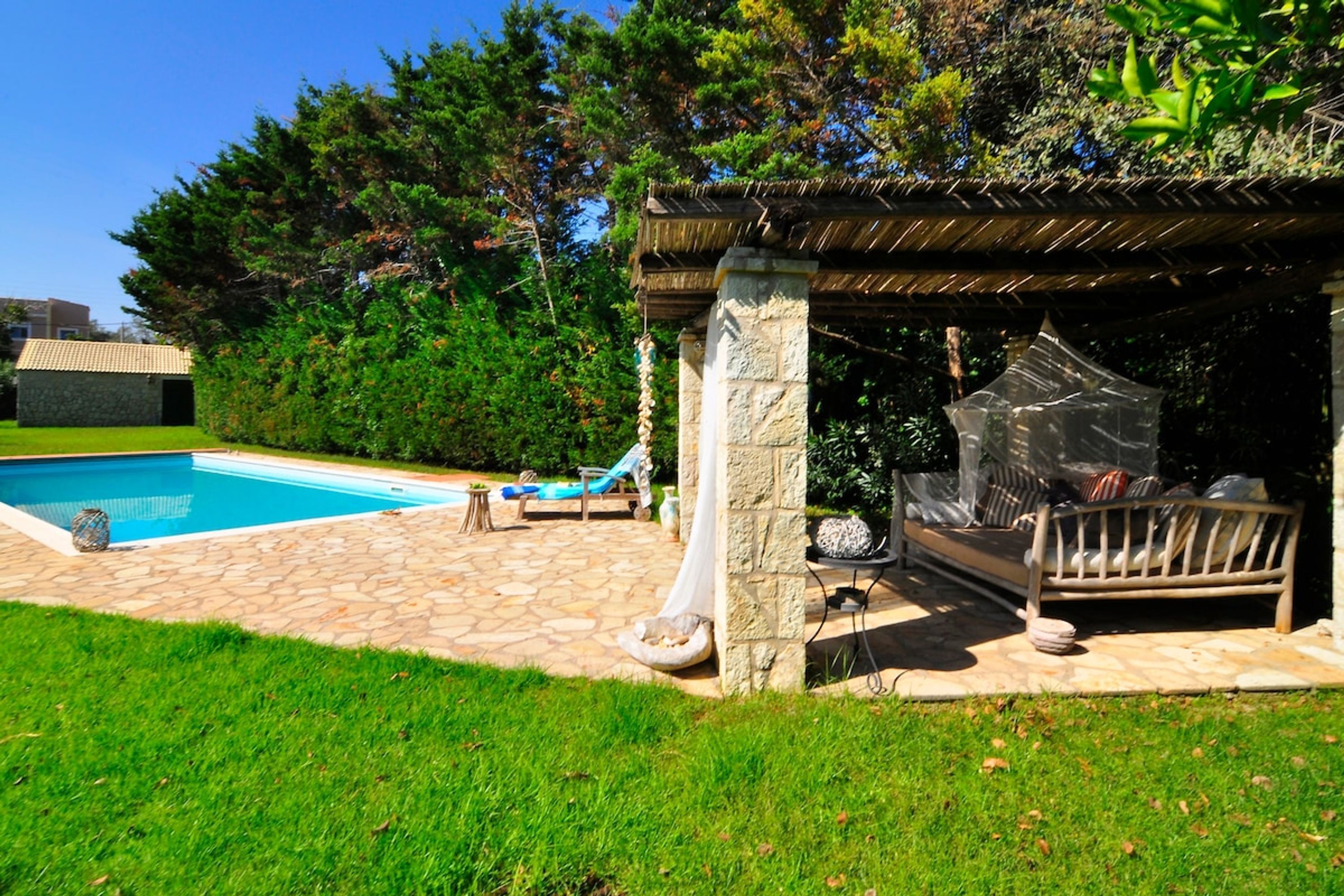 Un jardin avec une piscine, une terrasse en pierre, une terrasse en bois et un espace salon couvert.