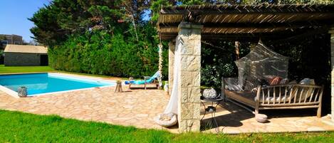 Un jardin avec une piscine, une terrasse en pierre, une terrasse en bois et un espace salon couvert.