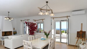A spacious living area with a white sofa, dining table, and chairs, a fireplace, and a view of the outdoors. - Dreamcatcher Sea View Mini Pool Paros (Paros)