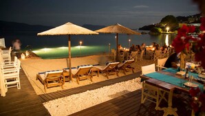 Un espace de restauration situé au bord de la plage, avec des tables et des chaises en bois, des parasols en chaume et une vue sur la mer la nuit.