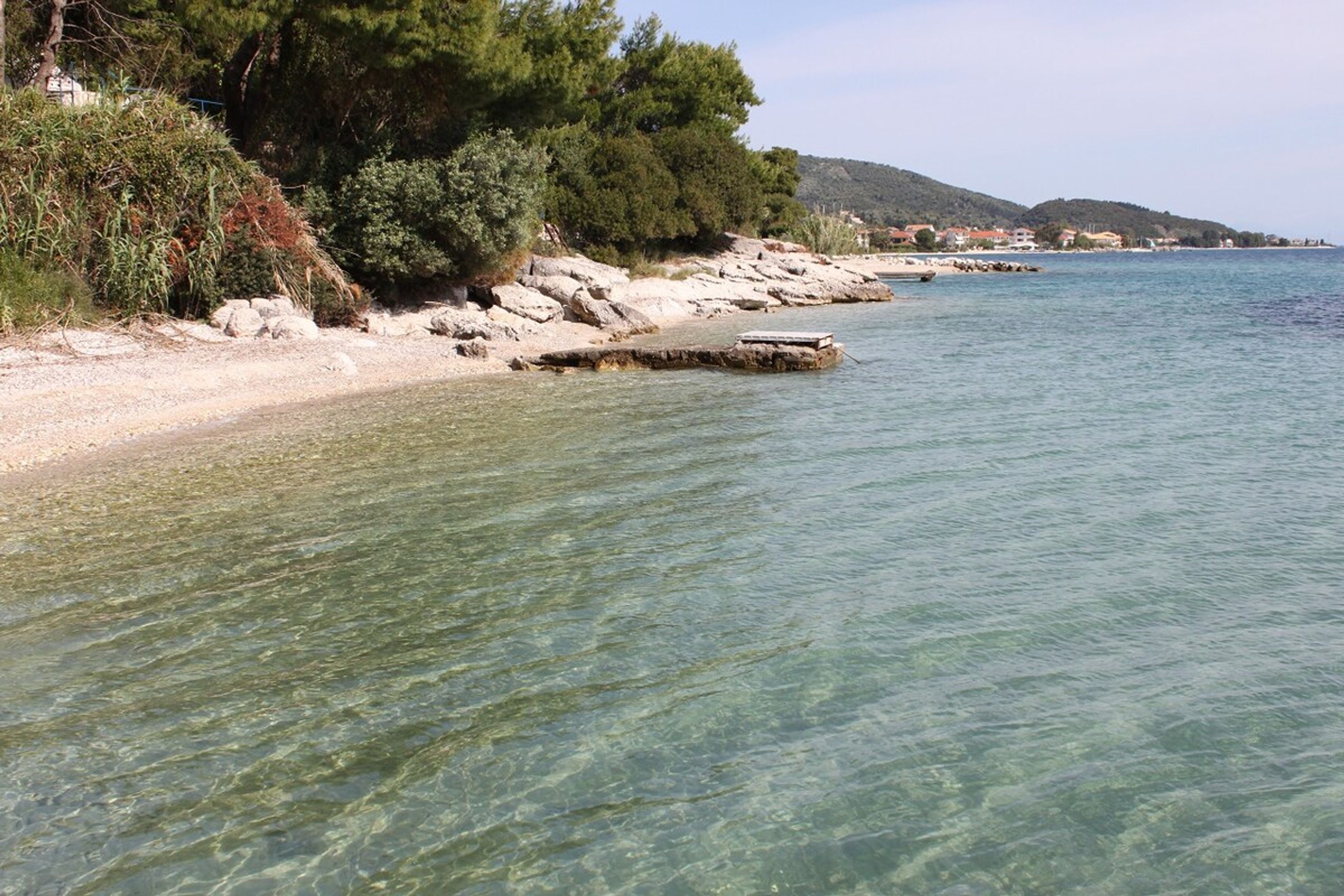A clear coastal view with rocky shoreline, clear water, and distant buildings.