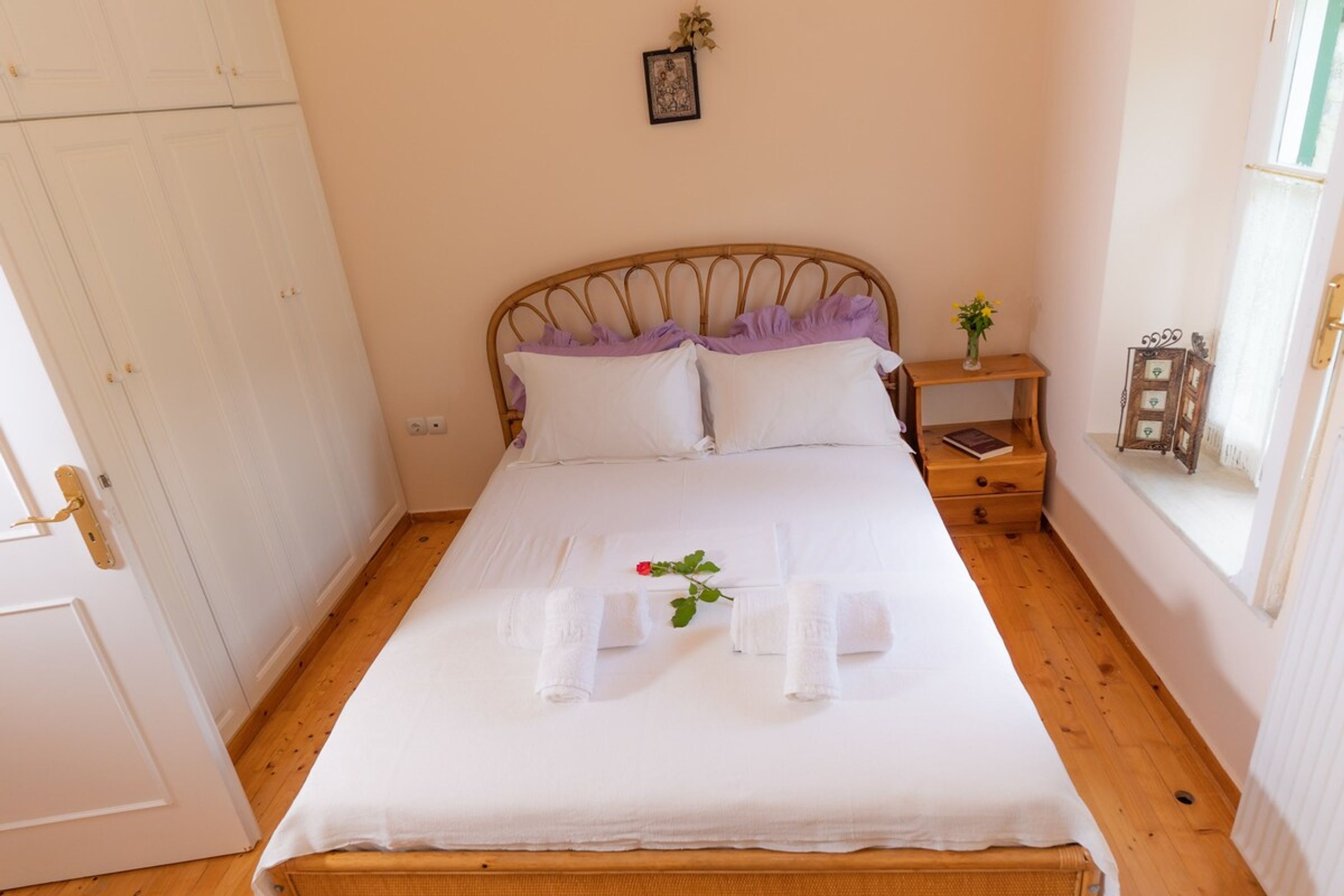 Cama bem arrumada com lençóis e toalhas brancas, cabeceira de madeira, mesa de cabeceira com um livro e um vaso de flores, e uma janela com vista para o exterior.