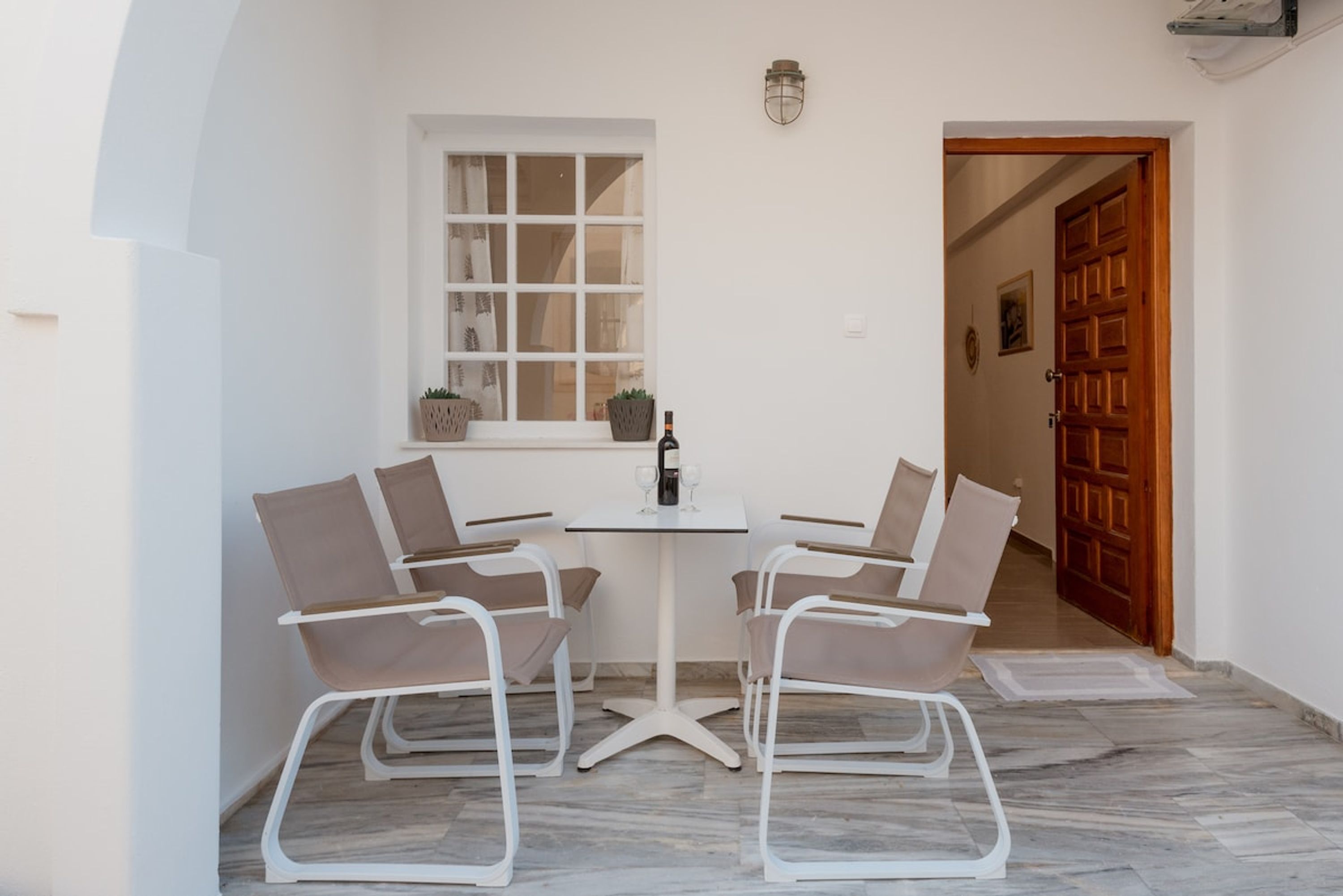 Un petit espace de restauration privatif, avec une table blanche et quatre chaises, une fenêtre avec des rideaux et une porte en bois en arrière-plan.