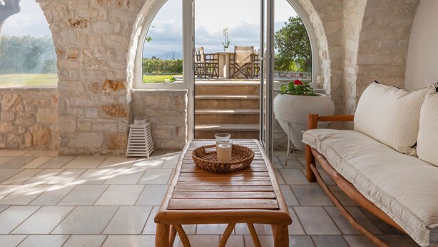 A stone-walled room with a wooden table, a cushioned bench, and a view of an outdoor terrace with a table and chairs. - Sunrise Serenity Near Ambelas Beach (Paros)