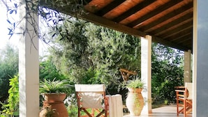 An outdoor covered patio with a wooden ceiling, large potted plants, a folding chair, and a tiled floor. - Agrilis Cottage Near the Beach (Trifylia)