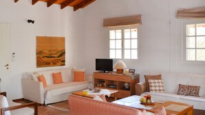 A cozy living room with a high wooden ceiling, a white sofa, a wooden cabinet with a television, and a window with a view. - Agrilis Cottage Near the Beach (Trifylia)