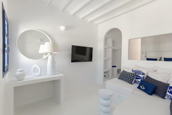 A modern, white-themed bedroom with a round mirror, a flat-screen TV, and a bed with patterned pillows. - Maria Sea View Apartment Naousa (Paros)