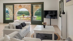 A living room with a white sofa, a coffee table, and a TV. There is a stone archway leading to an outdoor area with a table and chairs. - Villa Franca Ideal for Wedding Parties (Paros)