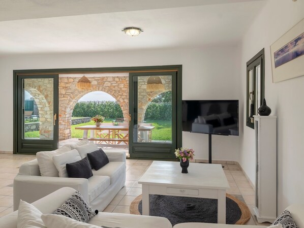 Una sala de estar con un sofá blanco, una mesa de centro y un televisor. Se observa un arco de piedra que da paso a un espacio al aire libre con una mesa y sillas.