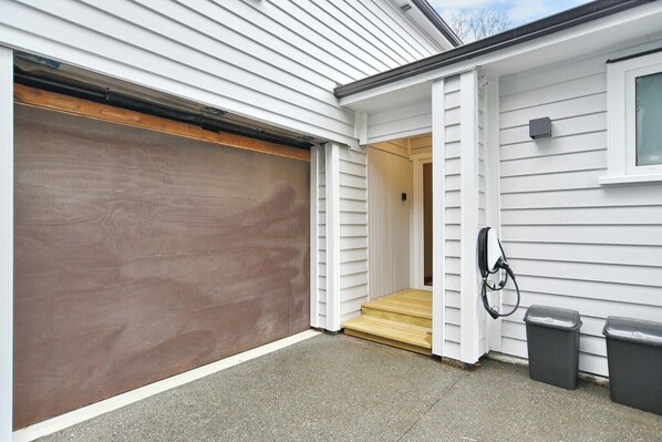 A garage with a closed brown door, a wooden step leading to a door, and two black trash bins beside the door.