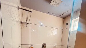A bathroom with white tiled walls, a black tiled floor, a glass shower enclosure, and a visible trash bin. - Spexxa Residency (Kozhikode)