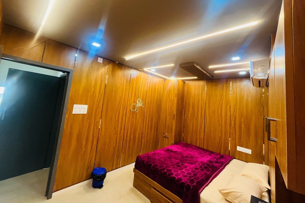 A bedroom with wooden paneling, a bed with a purple blanket, a blue trash bin, and a ceiling light fixture.