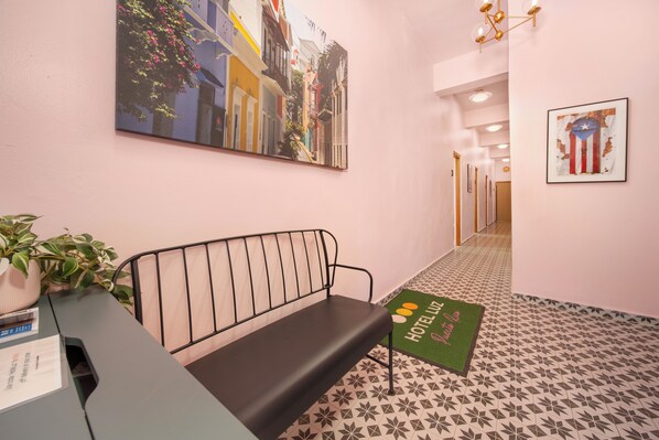 Vestíbulo de hotel con un banco, mostrador de recepción, felpudo verde con el texto 'HOTEL LUZ' y un cuadro grande de una escena callejera.