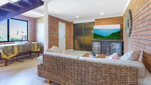 A living room with a wicker sofa, a dining table with chairs, a TV on the wall, and a brick wall.