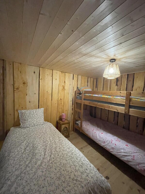 Une chambre dans un chalet en bois, avec un lit superposé, un lit simple et une petite table de chevet en bois avec une lampe.
