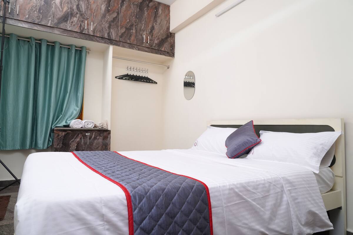 A neatly made bed with a quilted blanket, a pillow, and a headboard. There is a wooden cabinet with a mirror above it, and a towel rack with folded towels.