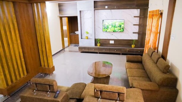 A modern living room with a sectional sofa, a coffee table, and a flat-screen TV mounted on a marble wall. - Sanskriti Homestay (Tirupati)