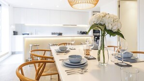 Un espace de repas moderne avec une longue table dressée pour un repas, un vase rempli de fleurs blanches et une cuisine en arrière-plan.