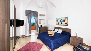 Een slaapkamer met een bed, nachtkastje, lamp, schilderij en uitzicht op zee door een raam.