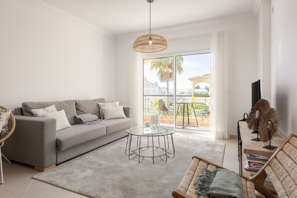 A modern living room with a grey sofa, a glass coffee table, wicker furniture, and a large window offering a view of the outdoors.