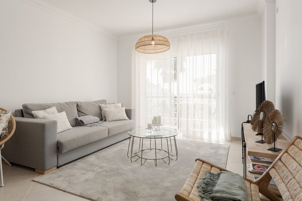 A modern living room with a grey sofa, a round glass coffee table, and a wicker chair. - Praia Village T2 - Peaceful condominium by HD (Vilamoura)