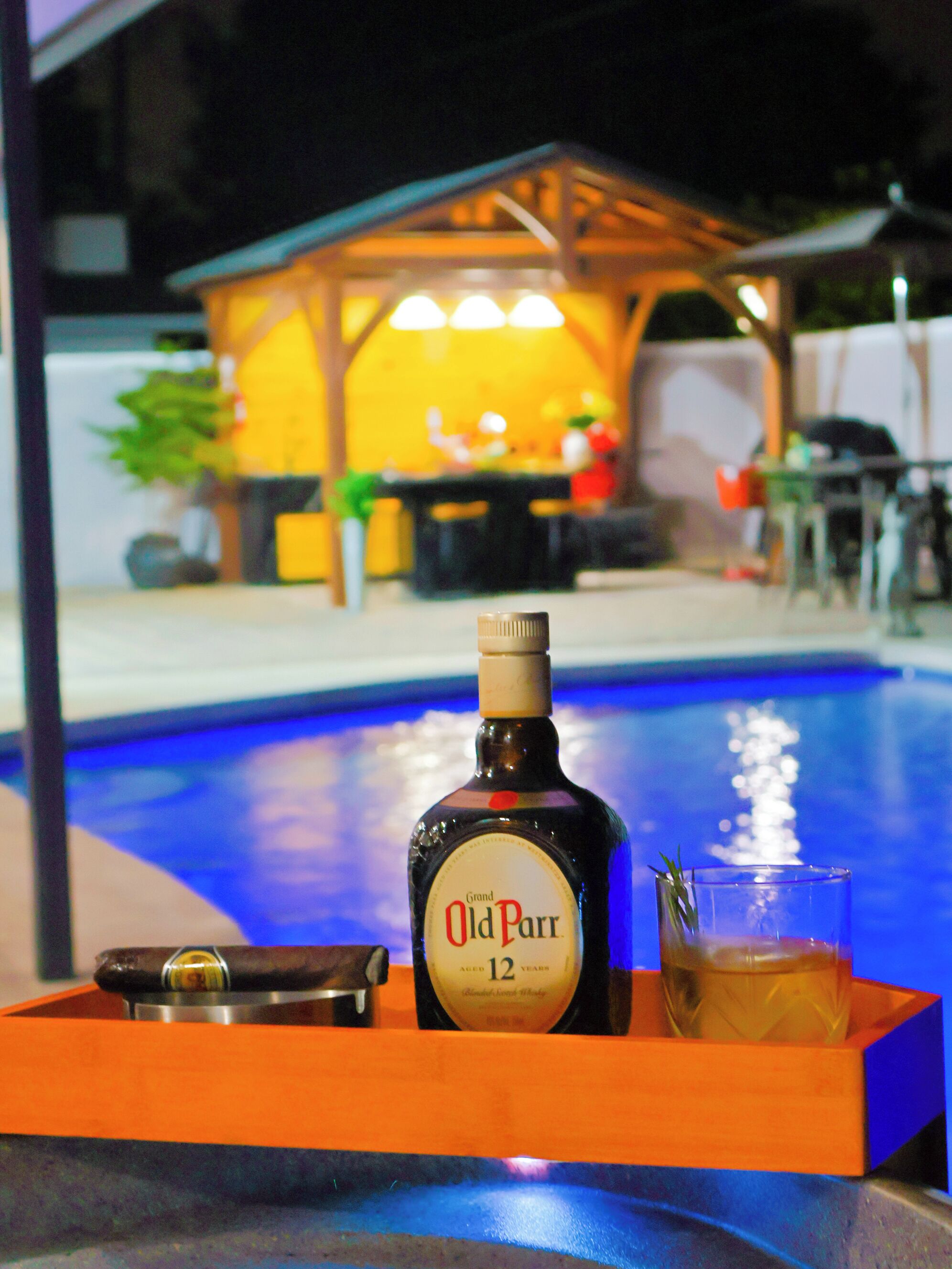Une bouteille de whisky Grand Old Parr, un verre contenant une boisson et un plateau placés au bord de la piscine.