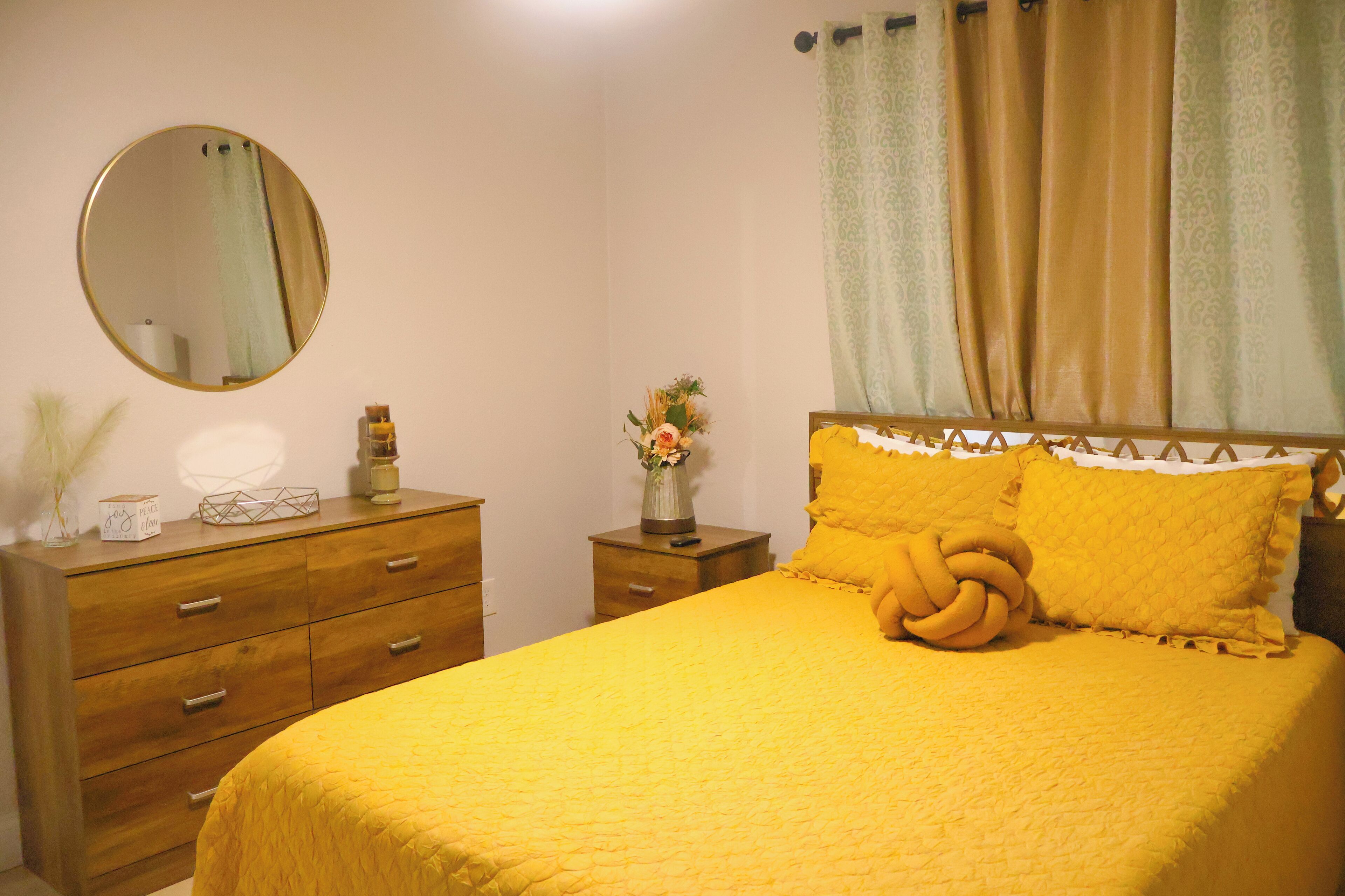A bedroom with a yellow bedspread, a wooden dresser, a mirror, a nightstand with a vase of flowers, and a window with curtains.