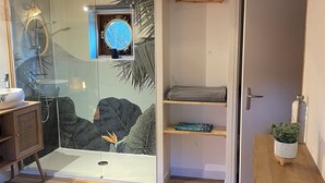 A modern bedroom with a wooden ceiling, a glass shower enclosure, a bed with a blue towel, a wooden dresser, and a small table with a plant. - Péniche Le Pescalune (Arles)