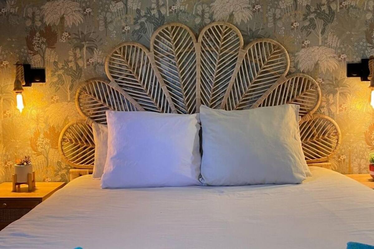 Un letto con una testiera dal motivo fogliare unico, due cuscini bianchi e un comodino con una pianta.