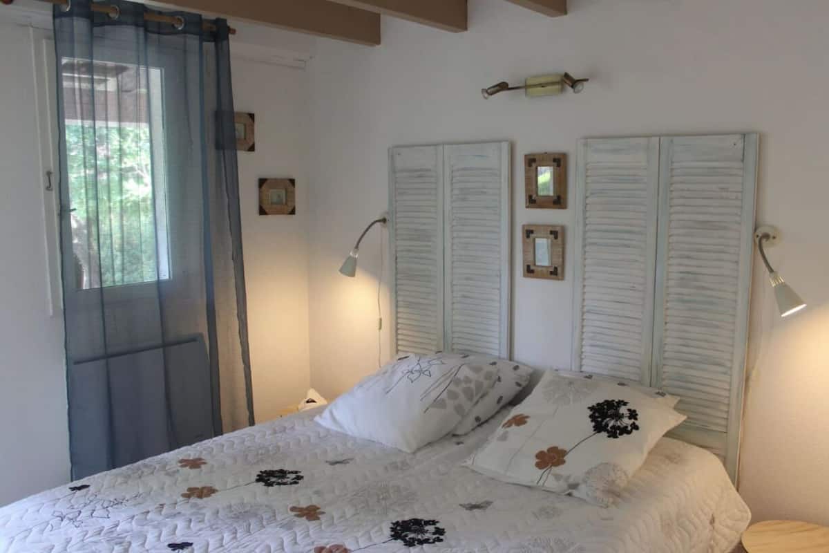 Um quarto com cama, cortinas brancas, janela e luminárias fixadas na parede.