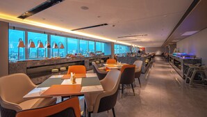 A modern restaurant with a long counter, orange chairs, and a city view through large windows. - Dengting Hotel (Haikou)