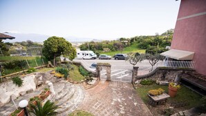 A driveway leading to a gated entrance with parked vehicles, a stone staircase, and a view of a green landscape with trees and distant mountains.