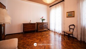 A room with a wooden parquet floor, a chandelier, a wooden cabinet with a plant, a chair, and a painting on the wall.