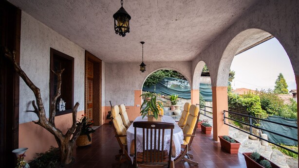 A covered patio area with a table and chairs, potted plants, and a view of the surrounding landscape.